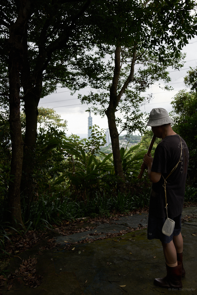A middle-aged Taiwanese man plays the flute far outside the city, the Taipei 101 in the background.