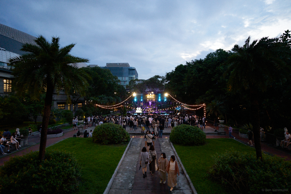 A stage surrounded by trees in the evening hours with a small crowd of people around it waiting for the band.