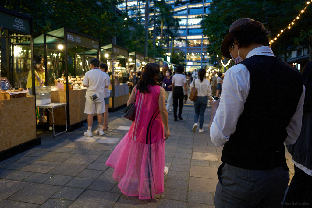 A lady in a vibrant pink dress walks alongside some food vendors, while a young man in a traditional suit stands in the forefront.
