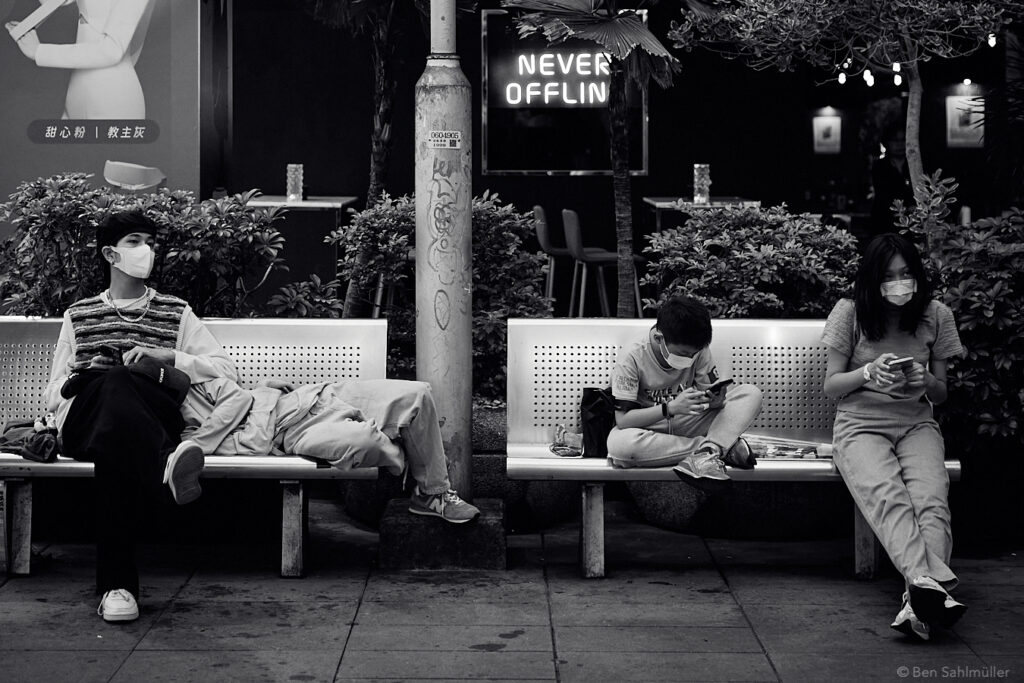 Four people on a bench before a sign saying "never offline." Three of them have their phones in hand.