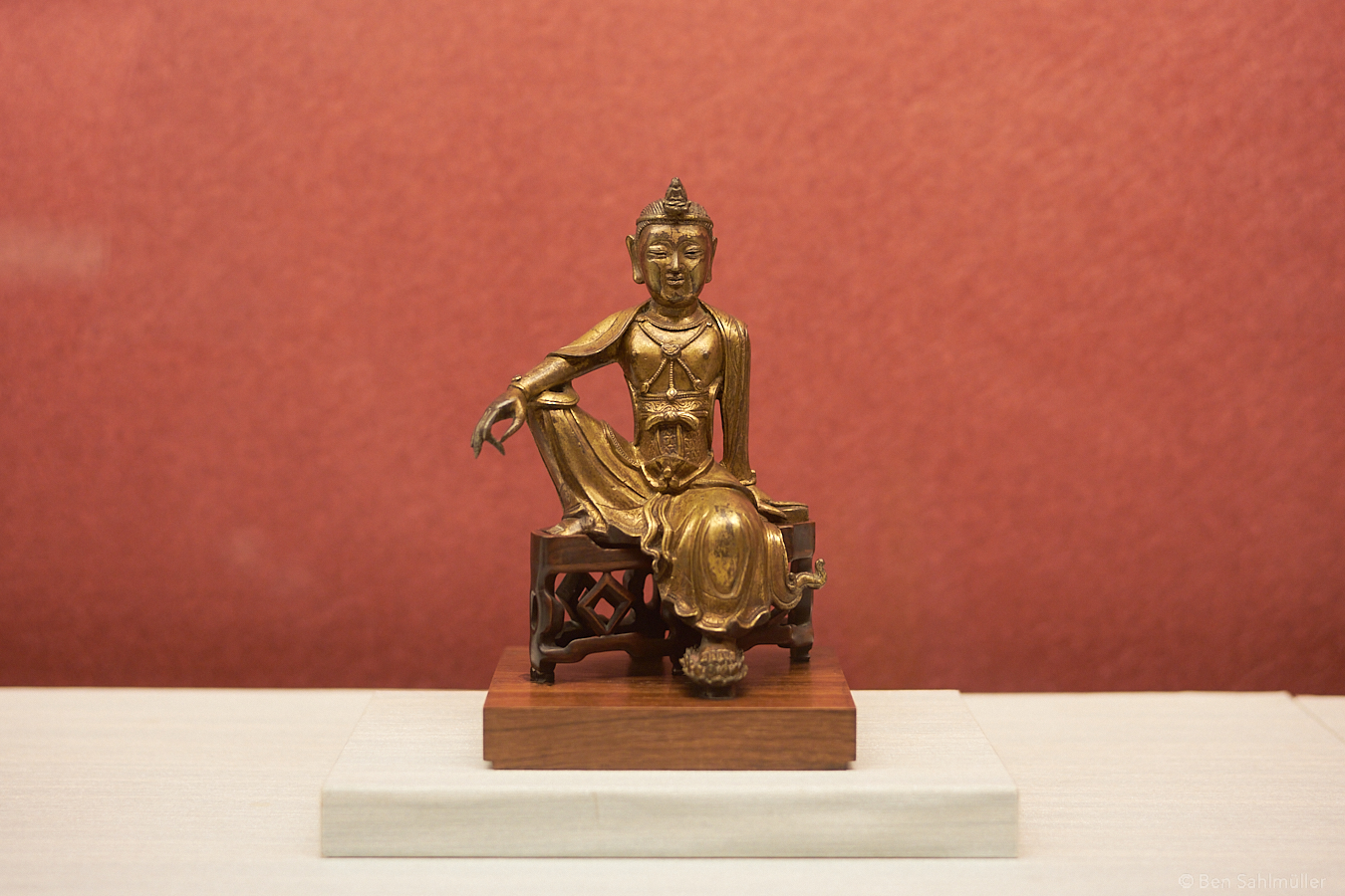A small statue of a monk. He is sitting on chair, with one arm at the side and the other stretched over his raised knees. He looks self-confident and filled with a peaceful joy.