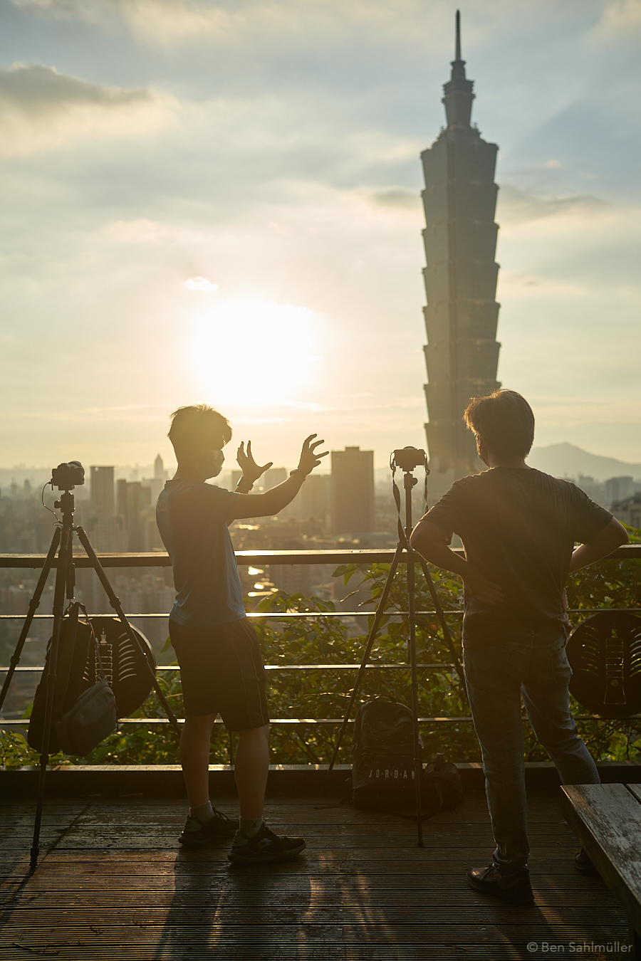Two photographers standing on a platform, the low sun and Taipei 101 in the background. The photographers have their cameras on tripod next to them and are discussing energetically, one of them gesturing with his hands.