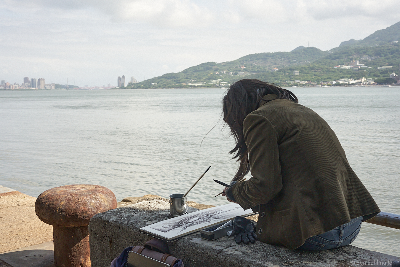 A woman sitting at the Tamsui River in Taipei, drawing the landscape with ink on a wide sheet of paper on the stone bench in front of her. She seems focused on her work and serene.