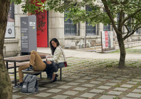 A couple on a bench, he is laying his head on her leg, she looks at him, while her hand caresses his hair.