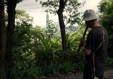 A Taiwanese man playing a flute in the woods. Taipei 101 can be seen in the background through some of the trees.