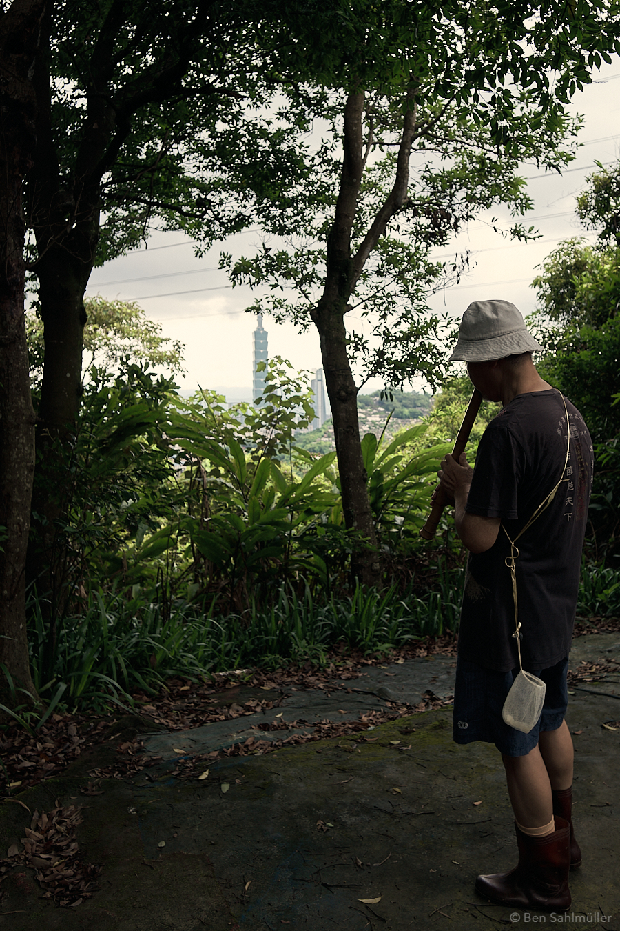 A Taiwanese man playing a flute in the woods. Taipei 101 can be seen in the background through some of the trees.