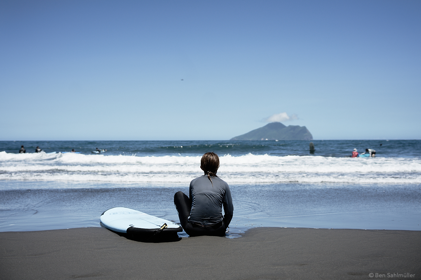 A women sitting at the waterfront on the beach. The sand has a dark grey, her board next to her has the color of the sea. She is mesmerized by the Ocean, looking out towards an island in the back.