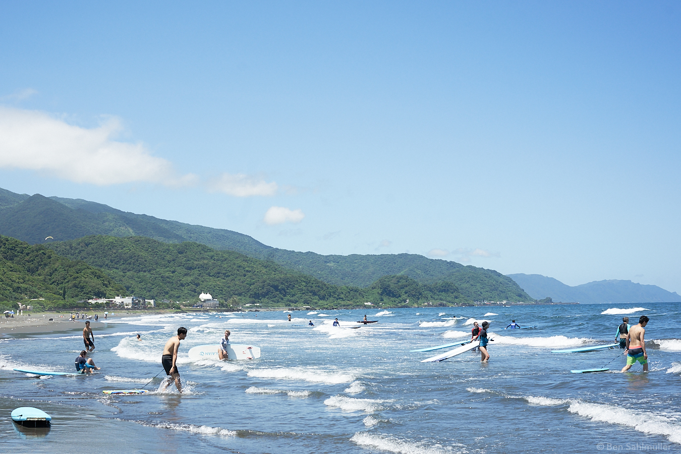 A beach, seen from the water's edge. There are many people tucking their surfboards into the waves. The weather is great with a clear blue sky.