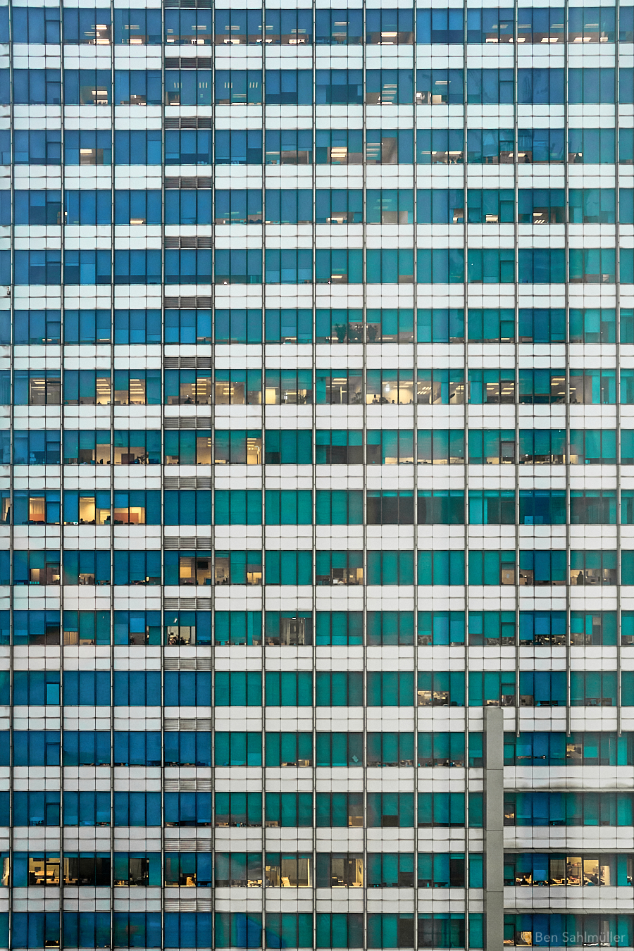 The side of an office building. A lot of glass and steel. Most of the blinds are closed, yet there is light behind the few that are still open. The offices behind look similar yet different in small ways. The silhouettes of people can be seen, all with similar jobs that are yet different in small ways.