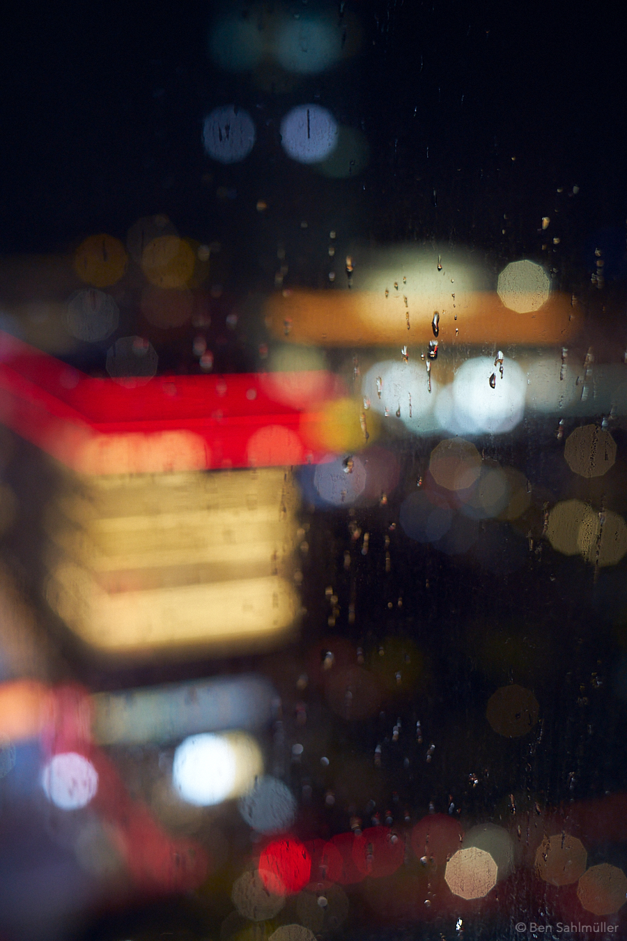 A few raindrops on a window by night, behind it the shape of a building can only be adumbrated by its shimmering lights in the window.