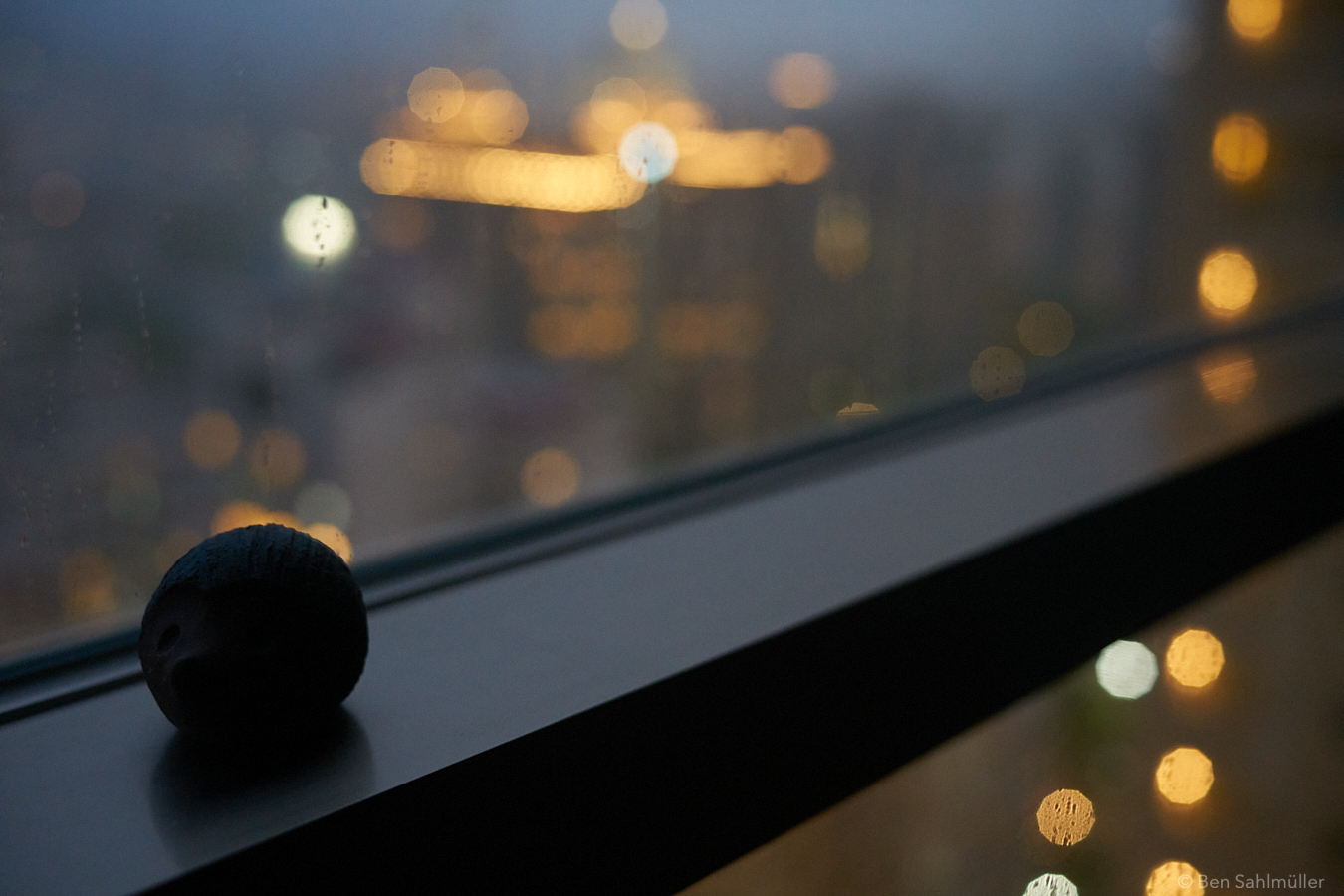 A clay hedgehog on a window sill, "looking" into the night outside. There are some buildings in the background, vaguely visible.