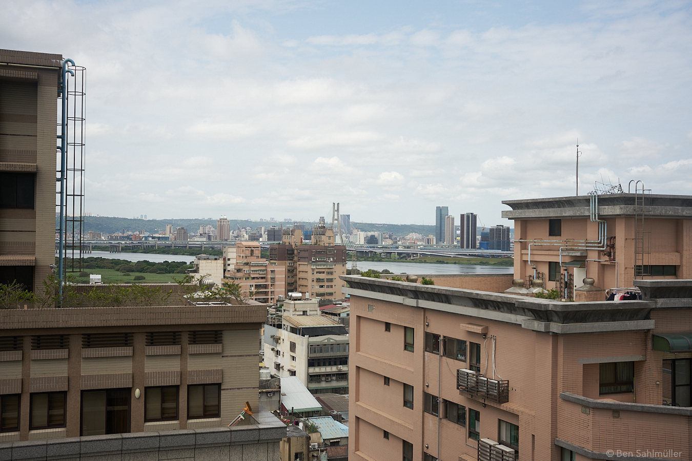 The view from a high rise. The sky is friendly and blue, there are lush mountain ranges in the distance and a river can be seen. Yet, all this is concealed by other high rises close to the window and feels out of reach.