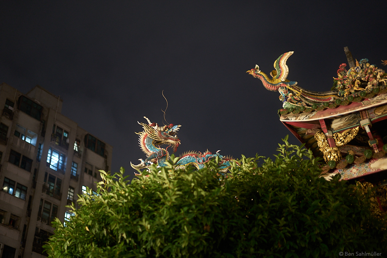 A dragon and a phoenix figure on a roof by night. The phoenix seems to take off while the dragon seems to growl at him. The warm light of the temple is contrasted by a cold glow from an apartment building in the back.