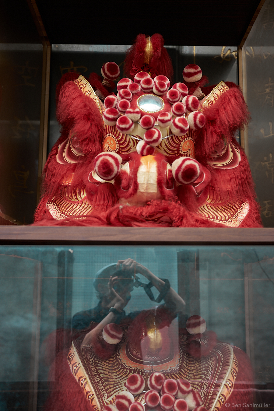 A red-golden mask used for a Chinese Lion Dance. In the glass pane underneath, a reflections shows me taking the photo.