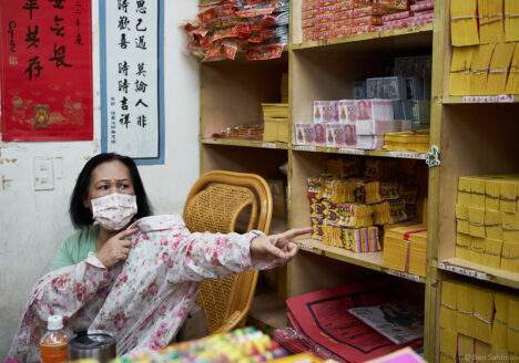 A woman shop owner shows us different kinds of joss paper in the piles of paper behind her desk.