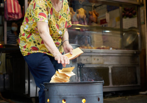 An older lady leaning forward to toss a pill of joss paper into an open fire. Her food stand can be seen in the back.
