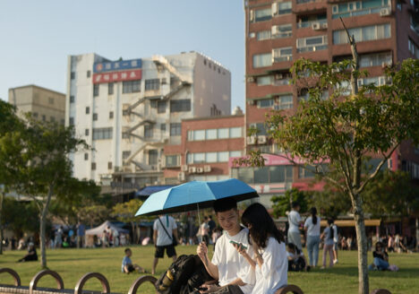 A young couple sitting on a bench at a park. He holds an umbrella to shield her from the sun while she is looking at her phone.