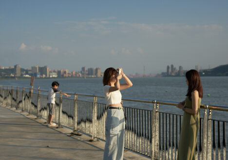 Two young women standing at a balustrade, one taking a photo of the other.