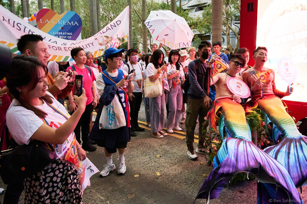 Two men dressed as rainbow mermen lasciviously lying in the red light of a pizza truck. Behind them a delegation of Australia and New Zealand holds a banner for LGBTQ rights. Several people are taking photos. Everyone is smiling.