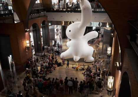 A giant (maybe 15m tall!) white rabbit in the open space of a mall. There is a performance artist standing in front of it, drawing a crowd of kids and families. The Rabbit has one arm raised as if preparing for a kung fu kick.
