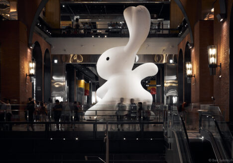 A giant (maybe 15m tall!) white rabbit in the open space of a mall. There is a performance artist standing in front of it, drawing a crowd of kids and families. The Rabbit has one arm raised as if preparing for a kung fu kick.