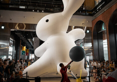 A giant (maybe 15m tall!) white rabbit in the open space of a mall. There is a performance artist standing in front of it, drawing a crowd of kids and families. The Rabbit has one arm raised as if preparing for a kung fu kick.