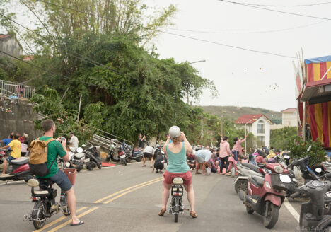 Two tourists sitting on bikes, taking photos as a crowd of people is catching candy thrown in high arcs from the stage.