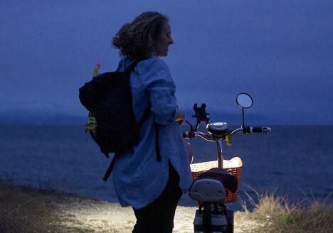 A young woman with a big backpack standing next to an electric moped at night.