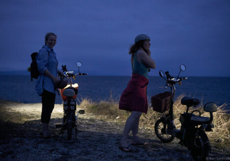Two young woman standing next to an electric mopeds at night. They are preparing their helmets and rides.