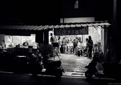 A night scene in an open diner, we are looking in from outside. The guests are looking happy, a woman is singing karaoke.