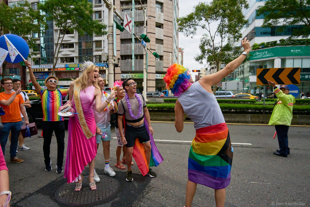A tall blond woman, wearing a shrill pink dress and a crown is taking a photo with a group of colorful participants of Taiwan Pride 2023.
