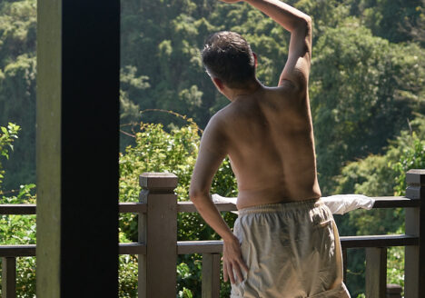 A man stretching while looking onto a forested hillside. He is wearing light shorts and no shirt.