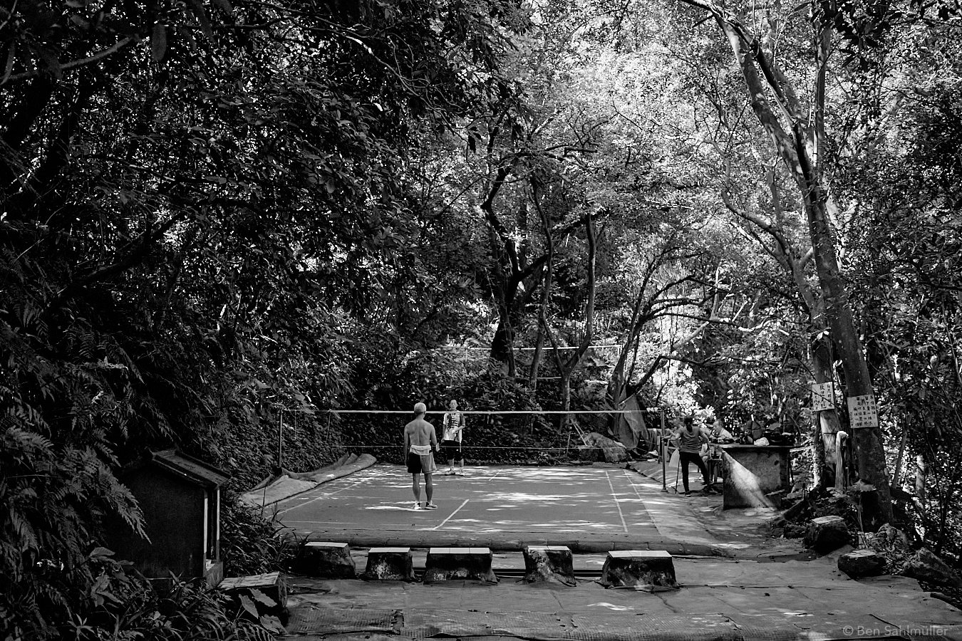 A group of older Taiwanese playing badminton. Their court is surrounded by a dense jungle forest of trees. Some of them are resting and chatting at the side. There is a small shrine to the left of the court.