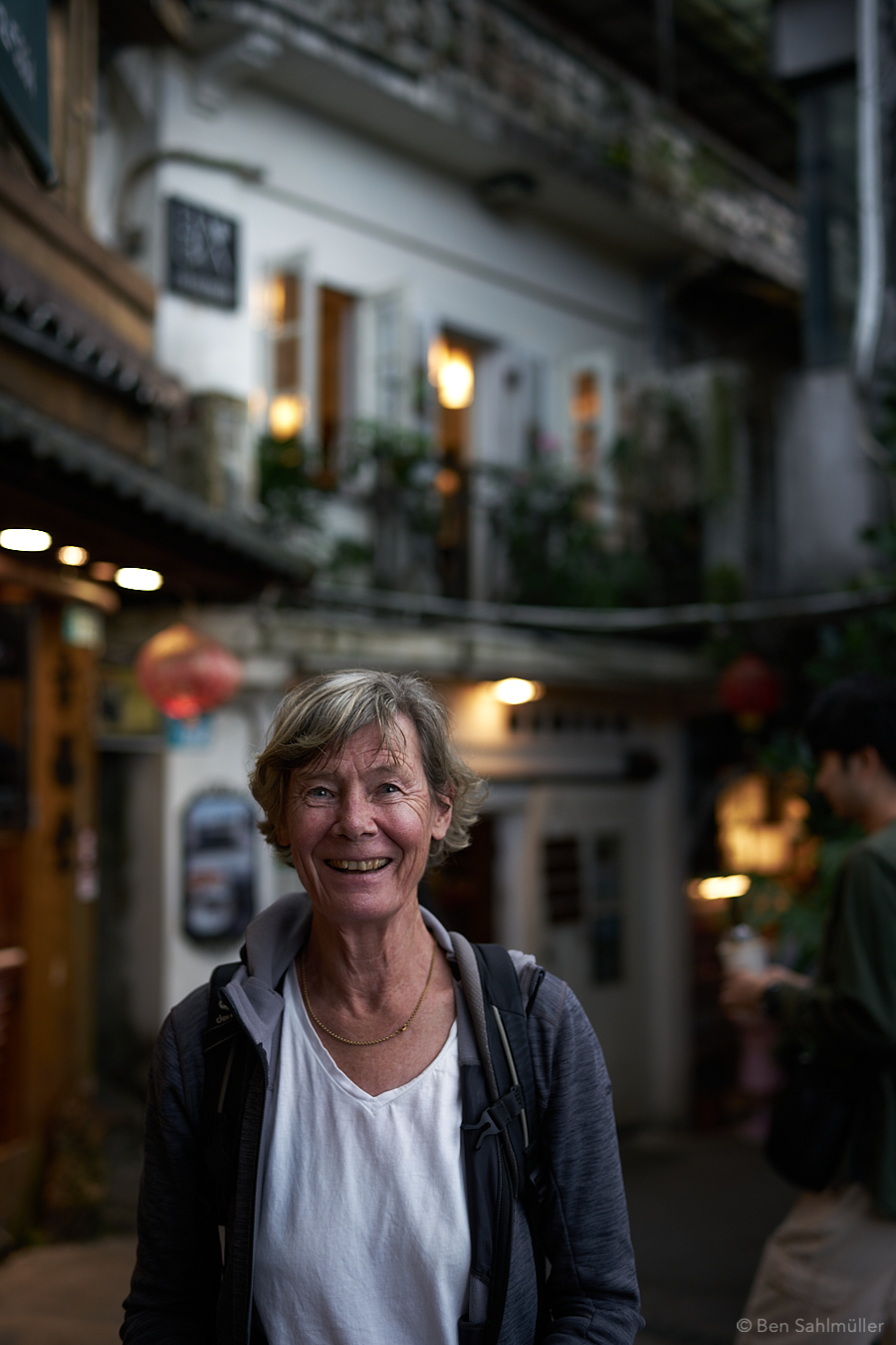 A portrait of a woman, wearing a white shirt and a grey jacket and smiling into the camera. The image is taken spontaneously in the streets of Jiufen. Some of the houses can be seen warm and blurred in the background.