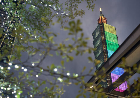 Taipei 101, as seen from a street below it. The branch of a tree seem to stretch towards it. There are warm fairy lights on the branch. At its north side, Taipei 101 is dressed by a giant LED screen which points towards us. The LEDs are lightened in rainbow colors.
