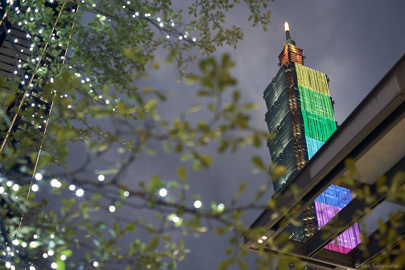 Taipei 101, as seen from a street below it. The branch of a tree seem to stretch towards it. There are warm fairy lights on the branch. At its north side, Taipei 101 is dressed by a giant LED screen which points towards us. The LEDs are lightened in rainbow colors.